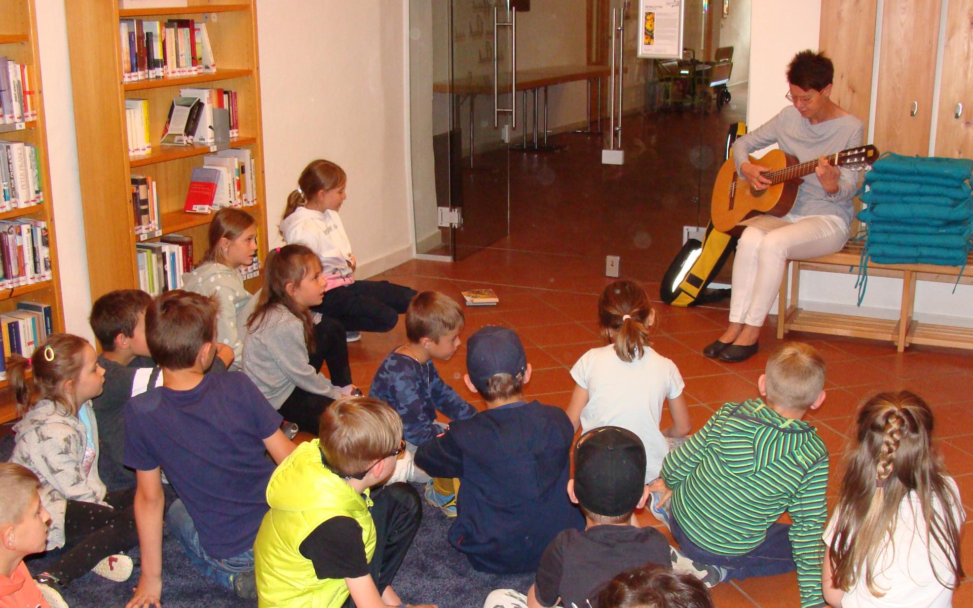 Frau mit Gitarre spielt sitzenden Kindern etwas vor.