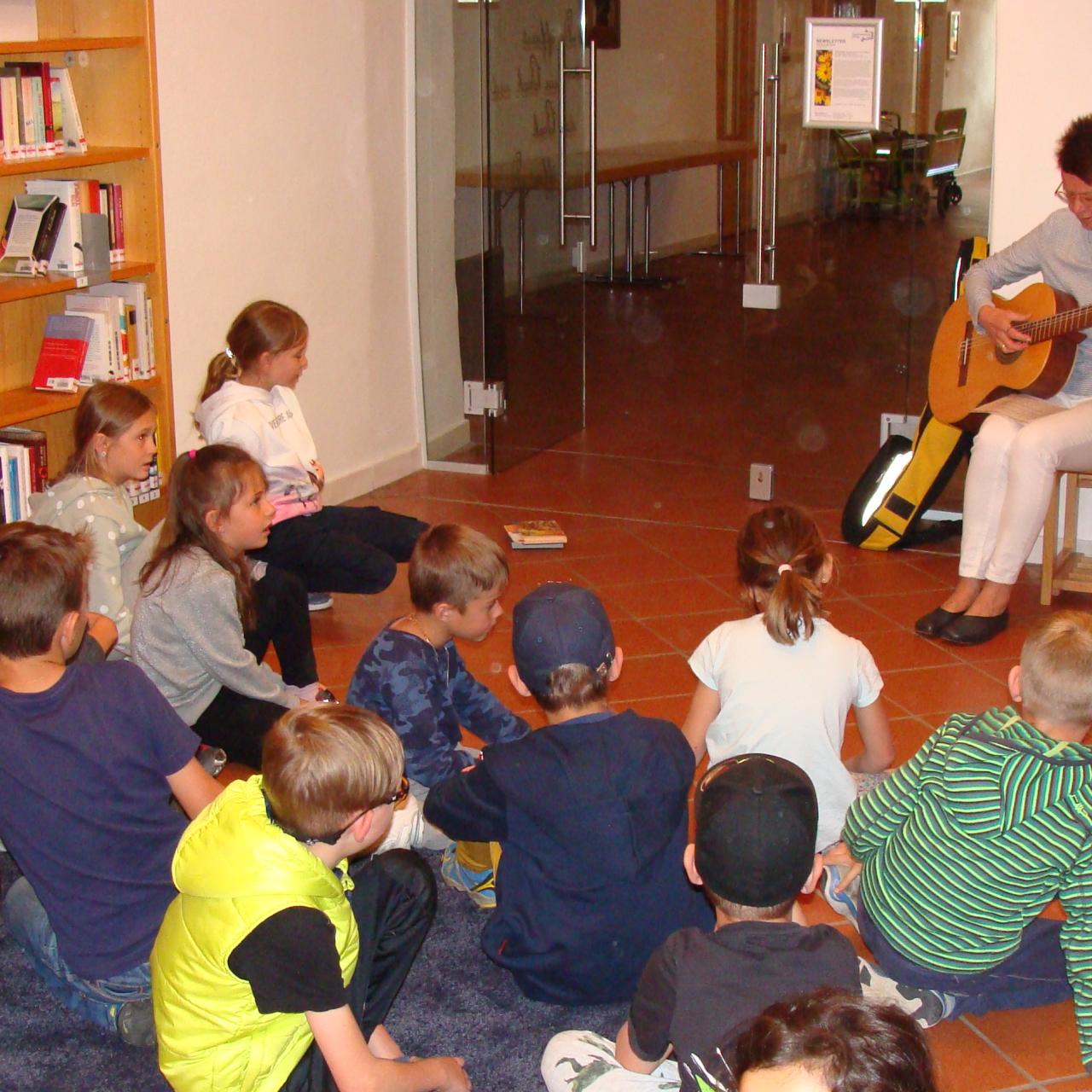 Frau mit Gitarre spielt sitzenden Kindern etwas vor.
